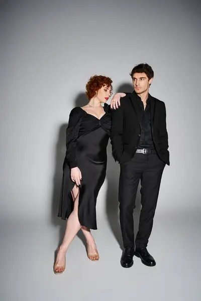Atractivo hombre en traje y mujer en vestido negro posando juntos sobre fondo gris, sexy pareja - foto de stock