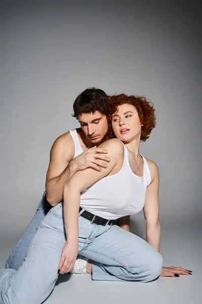 Atractivo hombre y mujer en pantalones vaqueros azules sentados en el suelo juntos y abrazando calurosamente, sexy pareja - foto de stock