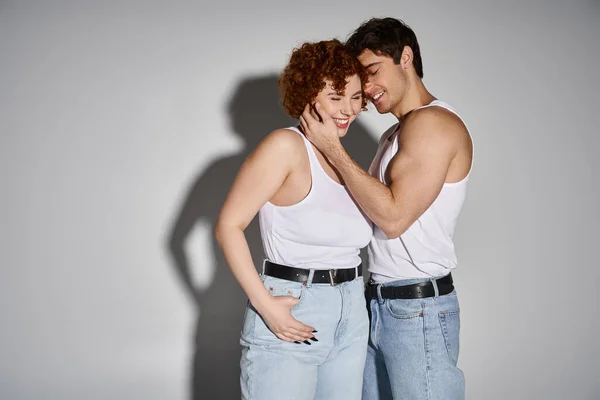 Cheerful alluring man and woman in jeans posing on gray backdrop and smiling happily, sexy couple — Stock Photo