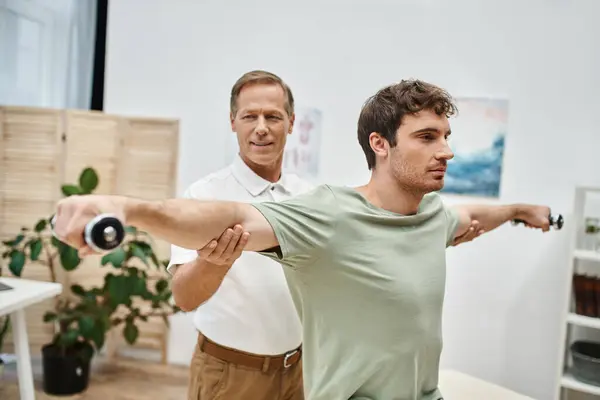 Cheerful mature doctor in uniform helping his handsome patient to use dumbbells on rehabilitation — Stock Photo