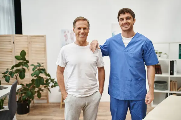 Joyeux beau docteur et son patient joyeux mature posant ensemble et souriant à la caméra — Photo de stock