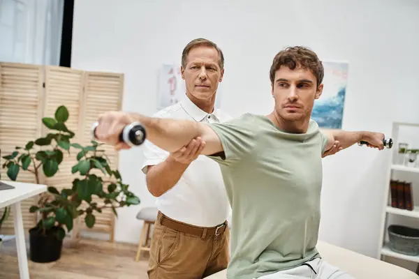 Attractive mature doctor helping his handsome patient to use dumbbells on rehabilitation in ward — Stock Photo