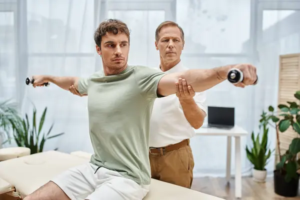 Hardworking mature doctor helping his handsome patient with dumbbells on rehabilitation in hospital — Stock Photo