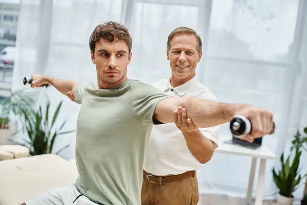 Dedicated jolly mature doctor helping his patient with dumbbells on rehabilitation in hospital — Stock Photo