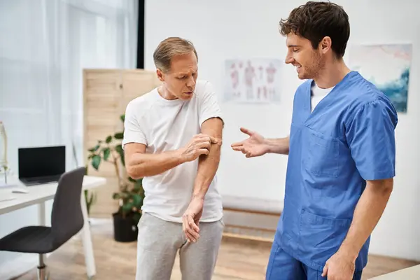 Bello gioioso medico in uniforme blu parlando con paziente maturo durante appuntamento, riabilitazione — Foto stock