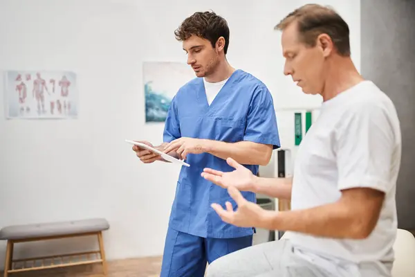 Handsome hardworking doctor holding tablet and listening to his mature patient, rehabilitation — Stock Photo