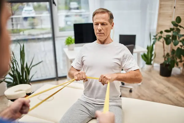 Fokus auf gutaussehendes Patiententraining mit Widerstandsband mit seinem verschwommenen Arzt, Rehabilitation — Stockfoto