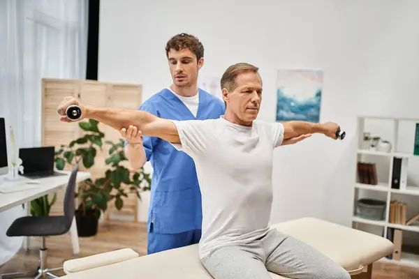 Handsome mature patient using dumbbells with help of his dedicated attractive doctor, rehabilitation — Stock Photo