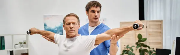 Attractive mature patient using dumbbells with help of his dedicated doctor, rehabilitation, banner — Stock Photo