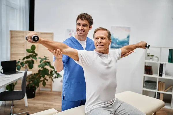 Cheerful mature patient using dumbbells during rehabilitation with help of his handsome jolly doctor — Stock Photo
