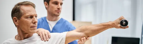 Médecin dévoué en robe bleue aidant patient attrayant mature avec haltère pendant la réadaptation — Photo de stock
