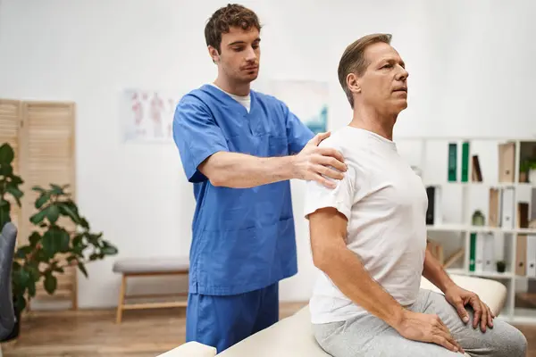 Gut aussehender Arzt in blauer Uniform hilft seinem reifen Patienten, seine Muskeln auf der Station zu rehabilitieren — Stockfoto