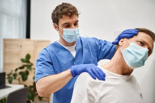 Beau médecin avec masque médical et gants aidant son patient à s'étirer pendant la réadaptation — Photo de stock