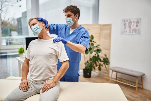 Devoted doctor with mask and gloves helping his patient to stretch his body, rehabilitation — Stock Photo