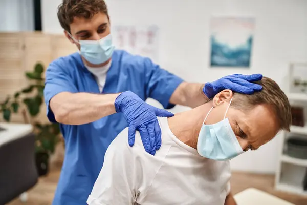 Gut aussehender Arzt mit Maske und Handschuhen, der seinen reifen Patienten während der Rehabilitation streckt — Stockfoto