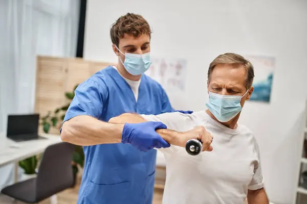 Médico trabalhador com máscara e luvas ajudando seu paciente a usar halteres durante a consulta — Fotografia de Stock
