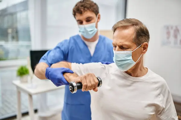 Devoted handsome doctor with mask and gloves helping his patient to use dumbbells on appointment — Stock Photo