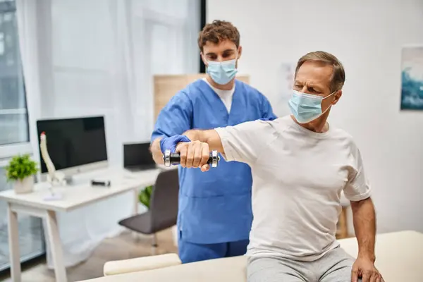 Engagierter Arzt in blauem Gewand mit Maske und Handschuhen, der dem Patienten hilft, die Hanteln in der Rehabilitation zu benutzen — Stockfoto