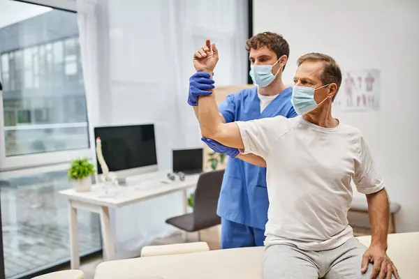 Engagierter Arzt mit Maske und Handschuhen, der seinem reifen Patienten hilft, seinen Körper auf der Station zu rehabilitieren — Stockfoto