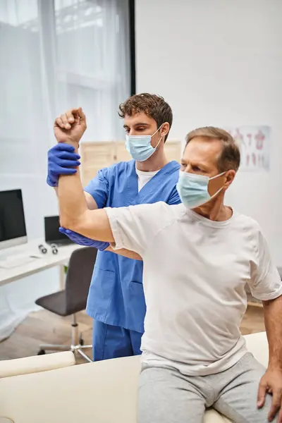 Hardworking doctor with mask and gloves helping mature patient to rehabilitate his muscles in ward — Stock Photo