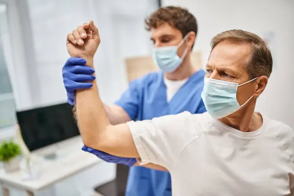 Médico dedicado com máscara médica e luvas ajudando paciente maduro para reabilitar seu corpo na enfermaria — Fotografia de Stock