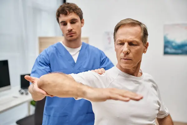 Good looking dedicated doctor in blue robe helping his patient to rehabilitate his body in ward — Stock Photo