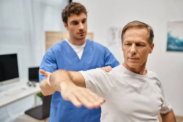 Attractive hardworking doctor in blue robe helping his patient to rehabilitate his body in ward — Stock Photo