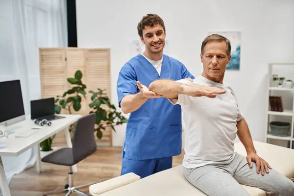 Cheerful good looking doctor in blue robe helping mature patient to stretch during rehabilitation — Stock Photo