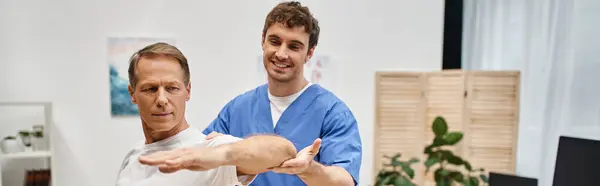 Cheerful handsome doctor helping his mature patient to stretch during rehabilitation, banner — Stock Photo