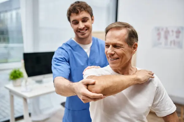 Jolly attractive doctor helping his mature patient to stretch during rehabilitation in hospital — Stock Photo