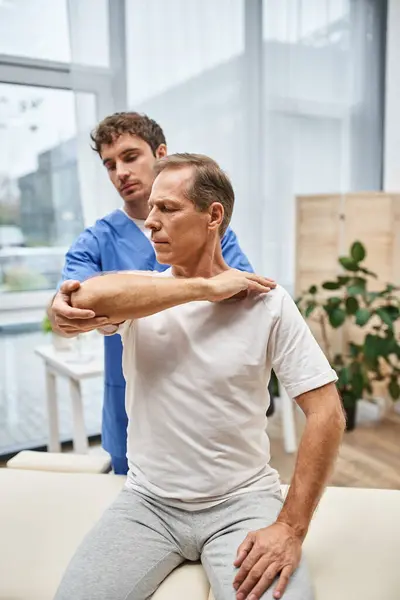Mature patient in casual attire stretching his muscles with help of his doctor, rehabilitation — Stock Photo