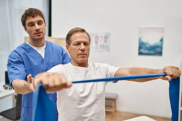 Beau médecin en uniforme bleu aidant son patient mature avec bande de résistance sur la réadaptation — Photo de stock
