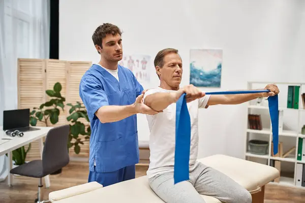 Attrayant médecin en robe bleue aider son patient mature avec bande de résistance sur la réhabilitation — Photo de stock