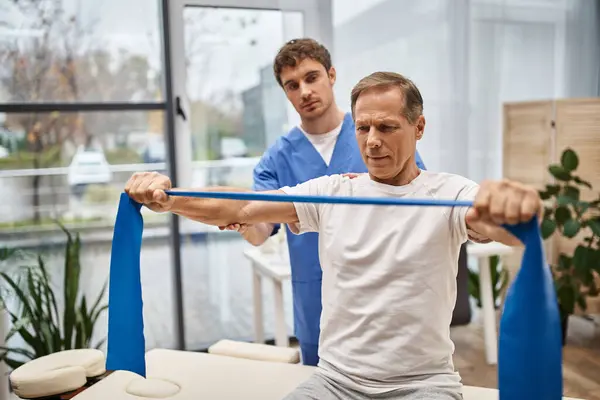 Fleißiger Arzt in blauer Uniform hilft seinem reifen Patienten mit Widerstandsband bei der Rehabilitation — Stockfoto