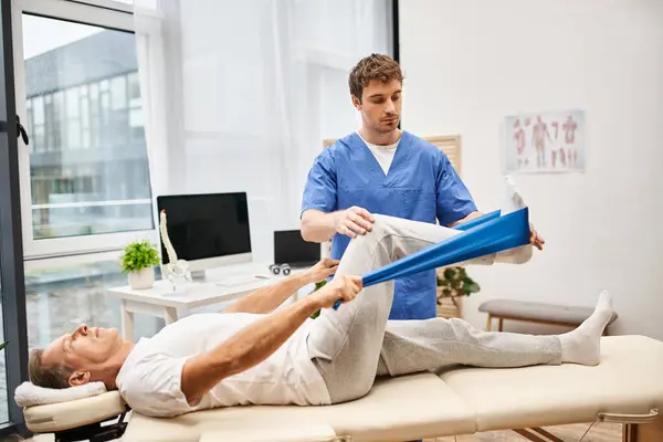 Good looking doctor in blue robe helping his handsome mature patient with resistance band in ward — Stock Photo
