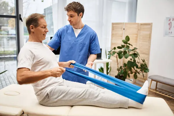 Appealing devoted doctor helping his mature joyous patient to use resistance band, rehabilitation — Stock Photo