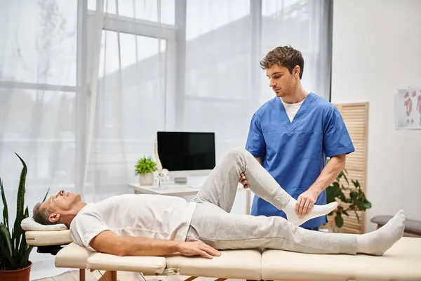 Attractive doctor in medical robe rehabilitating his mature handsome patient in hospital ward — Stock Photo