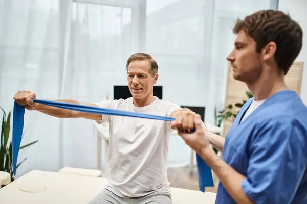 Handsome devoted doctor in blue robe helping his mature jolly patient to use resistance band — Stock Photo