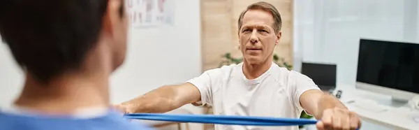 Focus on jolly handsome mature man using resistance band in front of his blurred doctor, banner — Stock Photo
