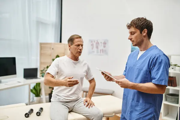 Good looking doctor in blue robe holding tablet during appointment with his mature handsome patient — Stock Photo