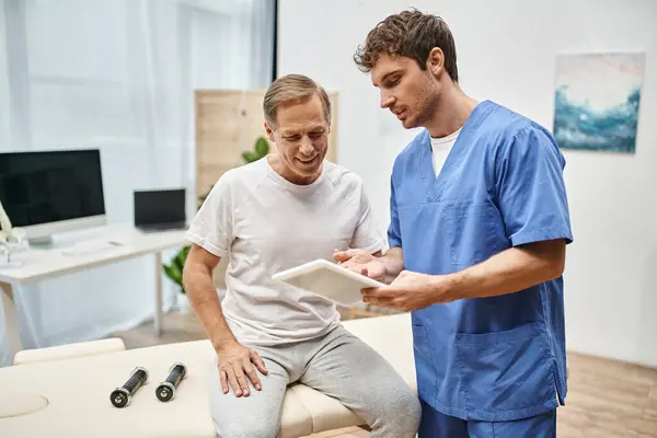 Attractive doctor in blue robe holding tablet and talking to his mature patient during appointment — Stock Photo