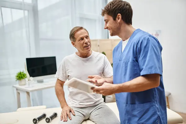 Happy devoted doctor holding tablet and talking to his mature jolly patient during rehabilitation — Stock Photo