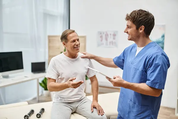 Lustiger engagierter Arzt hält Tablette in der Hand und spricht mit seinem reifen, fröhlichen Patienten während der Reha — Stockfoto