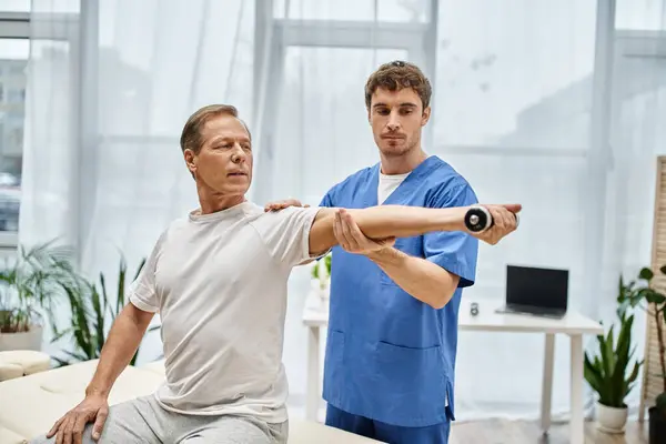 Guapo doctor en bata azul ayudando a su paciente maduro en traje casual a usar mancuerna en el hospital - foto de stock