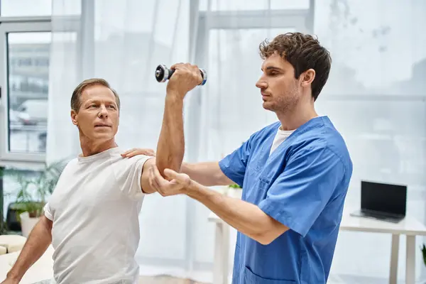 Hardworking doctor helping his mature patient in casual attire to use dumbbell on appointment — Stock Photo