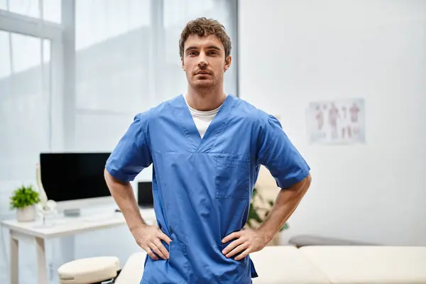 Good looking rehabilitologist in blue medical robe posing in hospital ward and looking at camera — Stock Photo