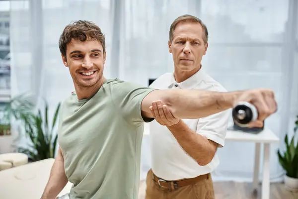 Good looking cheerful mature rehabilitologist helping his handsome patient to use dumbbell in ward — Stock Photo