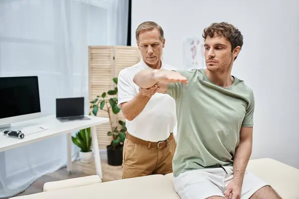 Good looking dedicated rehabilitologist helping his patient in casual attire to rehabilitate in ward — Stock Photo