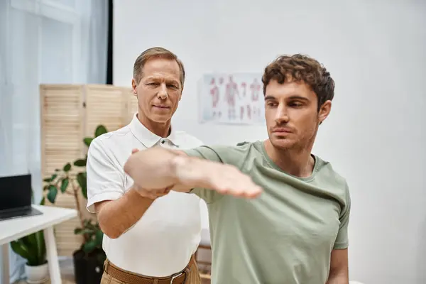 Attractive dedicated rehabilitologist helping his patient in casual outfit to rehabilitate in ward — Stock Photo