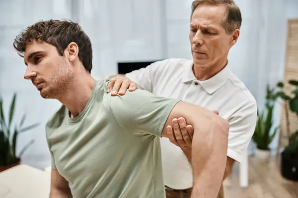 Good looking mature doctor in uniform helping his handsome patient to stretch during rehabilitation — Stock Photo